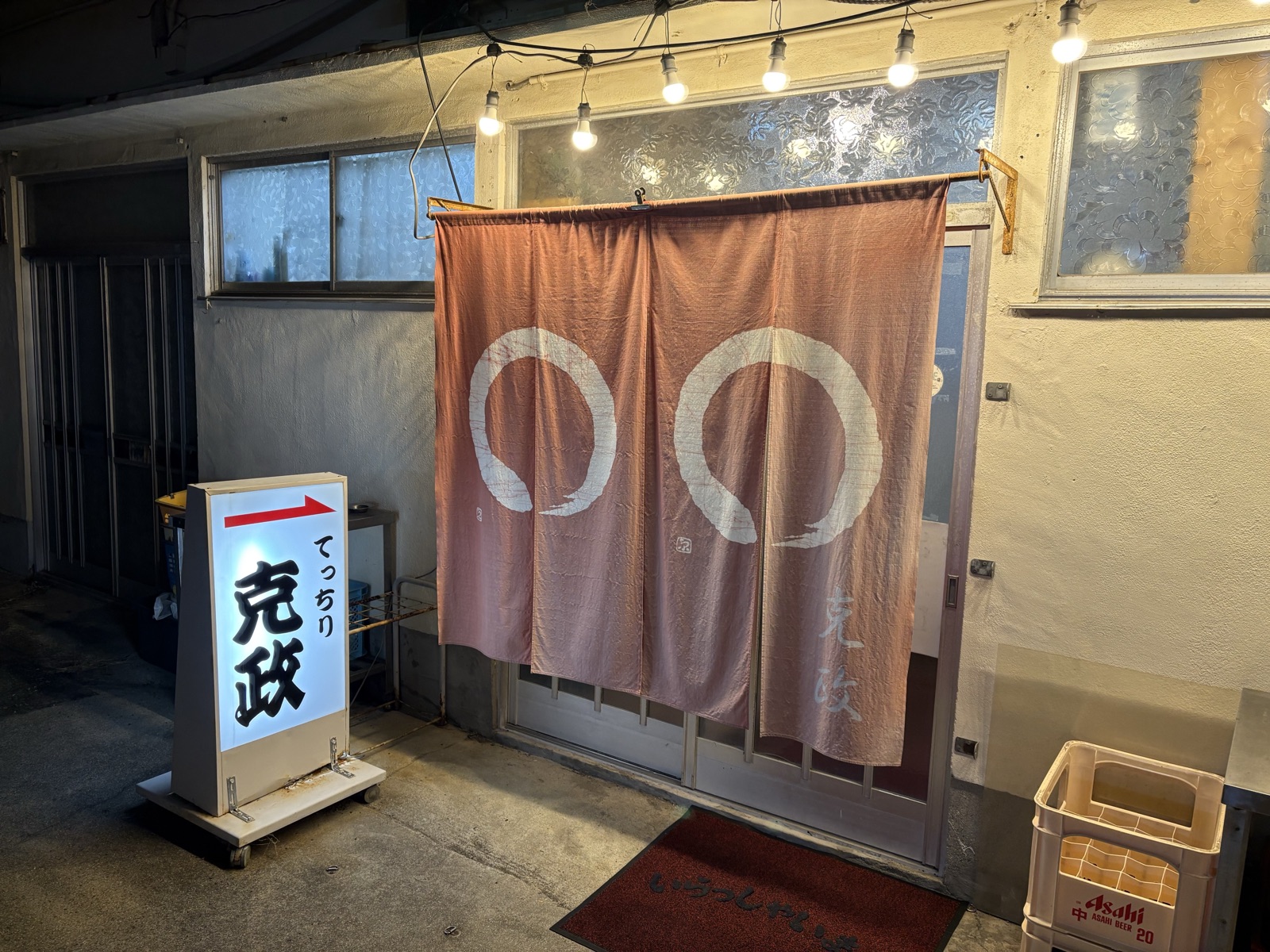 夜の店構え。裸電球の下に丸紋の暖簾が掛かり、傍らに「てっちり 克政」の行灯看板が立つ